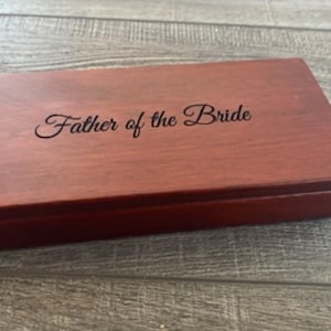 May include: A rectangular wooden box in a deep reddish-brown hue. The lid is inscribed with the words "Father of the Bride" in a cursive script. This box is likely intended as a commemorative gift or keepsake.