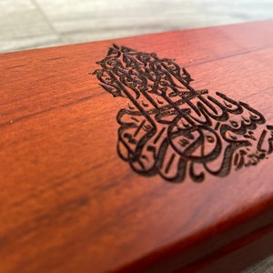 Könnte beinhalten: Eine Holzkiste mit dunkelbrauner Oberfläche. Die Kiste hat ein kunstvolles Design, das in die Oberseite geschnitzt ist und arabische Kalligraphie zeigt.