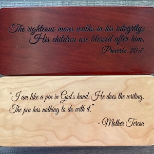 May include: Two wooden boxes with engraved text. The top box is a dark reddish-brown and reads "The righteous mans walks in his integrity: His children are blessed after him. Proverbs 20:7." The bottom box is light-coloured and reads "I am like a pen in God's hand. He does the writing. The pen has nothing to do with it." - Mother Teresa.