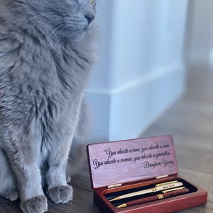 May include: A wooden pen box with two pens inside. The box is open, revealing the pens and an engraved quote. The quote reads, "You educate a man; you educate a man. You educate a woman; you educate a generation." A grey cat is in the background.