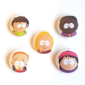 May include: Five circular pin-back badges featuring cartoon character portraits. The designs include a boy with brown hair in a green shirt, a boy with dark skin in a purple shirt, a character with blonde hair, a boy with orange hair, and a girl with black hair wearing a pink hat.