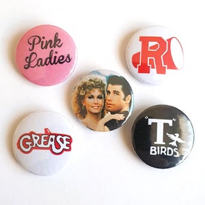 May include: Five circular pin badges with designs from the film Grease. One pink badge says "Pink Ladies". Others feature the word "Grease", a couple, the letter "R", and "T BIRDS" with a bird illustration.