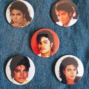 May include: Five round button badges featuring different images of Michael Jackson. The badges are white with a black border and are approximately 5 cm in diameter.