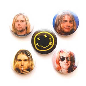 Könnte beinhalten: Ein Set aus fünf runden Ansteckbuttons. Vier Buttons zeigen fotografische Porträts einer Person mit schulterlangem blondem Haar in verschiedenen Posen. Der mittlere Button ist schwarz mit einer gelben, handgezeichneten Smiley-Grafik.