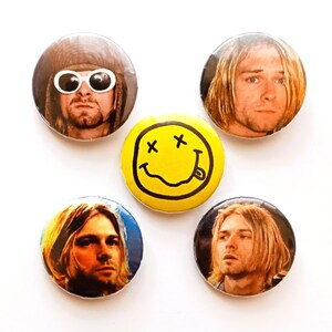 May include: A set of five circular pin-back badges. Four badges feature photographic portraits of a person with long blonde hair, one showing them in a brown hat and white sunglasses. The central badge is yellow with a black smiley face graphic.