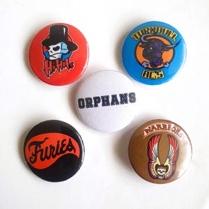 Puede incluir: Cinco botones redondos con diferentes diseños. Los botones son rojo, azul, blanco, negro y marrón. Los diseños incluyen una calavera con un sombrero de copa, un toro, la palabra "Orphans", la palabra "Furies" y una calavera con alas.