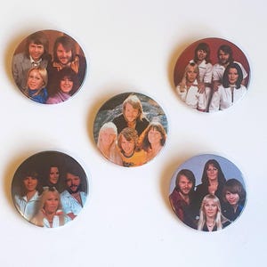 Puede incluir: Conjunto de siete botones redondos con pasador que representan a un grupo pop sueco. Los botones son blancos con un borde negro y presentan fotografías en color de los miembros del grupo.