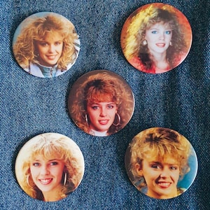 Puede incluir: Cinco botones redondos con alfiler que muestran a una mujer con cabello rubio. La mujer lleva una camisa blanca y tiene una sonrisa brillante. Los botones son de diferentes colores y tienen un acabado brillante.