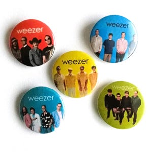 Può includere: Una collezione di cinque spille rotonde con la band Weezer. Ogni spilla ha uno sfondo di colore diverso: rosso, blu, giallo, verde acqua e verde. I membri della band sono raffigurati su ogni spilla.