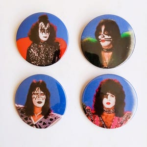 May include: Four round pinback buttons featuring the members of the rock band KISS. Each button has a blue background with a close-up image of a band member wearing their signature makeup.