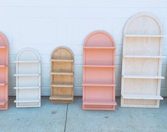 May include: Six arched wooden shelves with three shelves each. The shelves are painted in various colors, including pink, white, and natural wood.