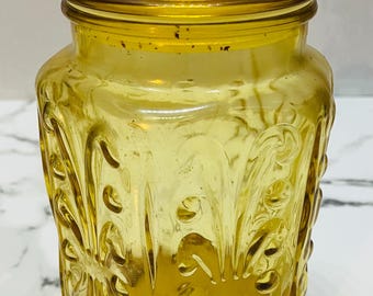 May include: A vintage yellow glass jar with a lid. The jar has a rounded shape with a decorative floral pattern embossed on the sides. The lid is also yellow and fits snugly on top. The jar is likely used for storage.