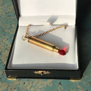 May include: A gold chain necklace with a bullet casing pendant. The bullet casing is topped with a red gemstone.