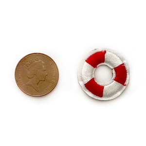 Puede incluir: Un parche bordado con forma de salvavidas, con segmentos rojos y blancos. Junto a él, una moneda. El parche mide aproximadamente 2,5 cm de diámetro. Ideal para manualidades de temática náutica.