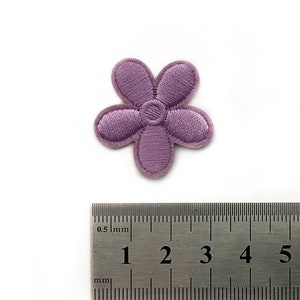 Puede incluir: Un parche bordado en forma de flor de color morado, que mide aproximadamente 2,5 cm de diámetro, se muestra junto a una regla.