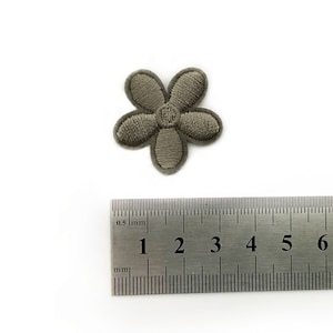 Op de afbeelding: Een klein grijs geborduurd bloemenpatch, ongeveer 1,5 cm in diameter. De bloem heeft vijf bloemblaadjes en een klein midden.