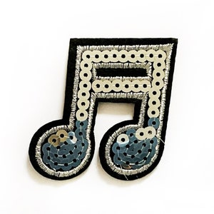 May include: A black, sequined musical note patch. The patch features a black felt base with silver and blue sequins. A ruler is placed below the patch for size reference.