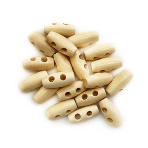 May include: A collection of light beige wooden toggle buttons, each featuring a cylindrical shape with two centered holes for attachment. These small fasteners have a smooth, natural finish and are arranged in a loose, overlapping pile.