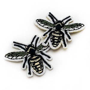 May include: Three embroidered bee patches. Each patch features a black and gold striped body, black legs and antennae, and green and white wings. The patches are on a white background.