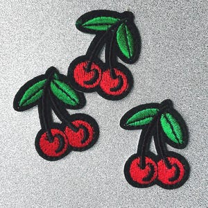 Op de afbeelding: Drie geborduurde patches met rode kersen, groene bladeren en zwarte stelen. De patches liggen op een zilveren, getextureerde achtergrond. Deze patches zijn geschikt om kleding of accessoires te versieren.