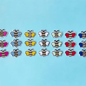 May include: Embroidered bee patches in a range of colours, including pink, blue, yellow, white, red, and purple, set against a light blue backdrop. Each patch depicts a black and tan bee with white wings.
