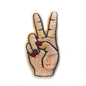 May include: A beige embroidered peace sign patch with red nail polish on the fingers.