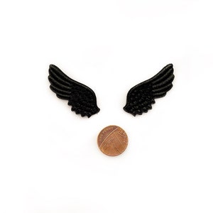 May include: Two black felt wings, each about the size of a US penny, are arranged symmetrically on a white background.