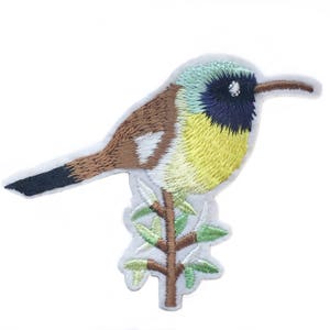 以下が含まれることがあります： 緑の枝に止まっている黄色と青の鳥の刺繍パッチ。鳥は黒い目をしており、体は茶色です。