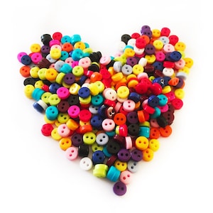 Può includere: Una composizione a forma di cuore di bottoni colorati in varie tonalità, tra cui rosso, giallo, blu, verde e viola. Ogni bottone ha due fori ed è realizzato in un materiale liscio e lucido. I bottoni sono sparsi su uno sfondo bianco.