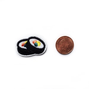 May include: Two embroidered sushi rolls, one on top of the other, with a black outline and colorful filling. The sushi rolls are about the size of a British one pence coin.