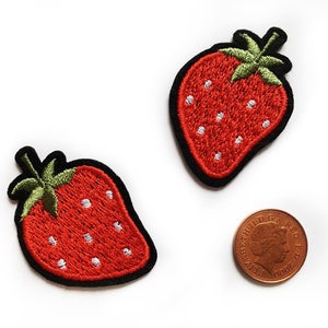 以下が含まれることがあります： 赤い果実、緑の葉、白い種が付いた、2つの刺繍されたイチゴのパッチ。パッチには黒い縁取りがあります。サイズの比較のために、パッチの隣にペニーがあります。白い背景にあります。
