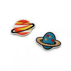 May include: Two embroidered iron-on patches of planets with rings. The first patch is orange and yellow with a white ring. The second patch is blue with red spots and a beige ring.