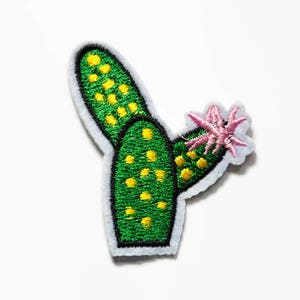 Puede incluir: Un parche bordado termoadhesivo con forma de cactus verde con manchas amarillas y una flor rosa floreciendo en el lateral. El cactus tiene un contorno negro grueso y un borde de fieltro blanco, con costuras detalladas en todo el diseño.