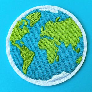 Op de afbeelding: Geborduurde patch met een gestileerde wereldkaart. De continenten zijn heldergroen tegen een blauwe oceaanachtergrond. De patch heeft een witte rand en is cirkelvormig.
