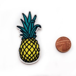 Puede incluir: Un parche bordado de piña con cuerpo amarillo, sombreado cruzado negro y hojas verde azulado. El parche está colocado junto a una moneda británica de un penique para comparar el tamaño, con un diámetro de aproximadamente 2,03 cm.