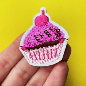 May include: A cupcake-shaped patch with pink sequins, brown frosting, and a pink embroidered wrapper. The patch has a white felt border and a pink cherry on top. The background is yellow.