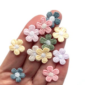 May include: A collection of small, embroidered flower appliques in various pastel colors, including pink, yellow, green, and blue. Each flower has a white center and is approximately 1 inch in diameter. These decorative appliques are ideal for craft projects.