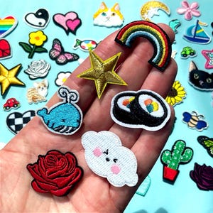 Könnte beinhalten: Eine Sammlung bunter, gestickter Aufnäher in verschiedenen Formen, darunter ein Regenbogen, ein Stern, Sushi-Rollen, ein Wal, eine Rose und eine Wolke mit Gesicht. Die Aufnäher befinden sich auf hellblauem Hintergrund.