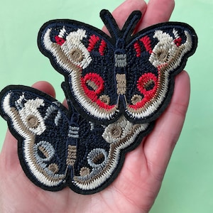 May include: Two embroidered butterfly patches. The butterflies have navy blue wings with red, white, and tan details. The patches are held in a hand against a light green background.