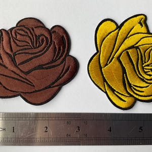 Puede incluir: Dos parches bordados de rosas. Uno es marrón con contorno negro, el otro es amarillo con contorno negro. Una regla debajo muestra las medidas en centímetros.
