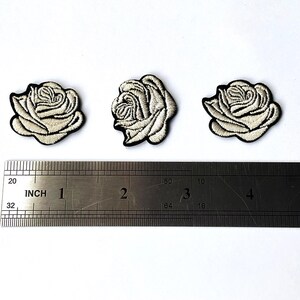 Puede incluir: Tres parches bordados de rosas con pétalos color crema y contornos negros. Una regla metálica con medidas en centímetros se coloca debajo. Cada parche tiene aproximadamente 3,8 cm de diámetro.