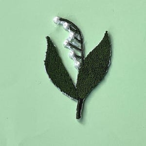 以下が含まれることがあります： スズランの刺繍パッチ。濃い緑色の葉と白いベル型の花が特徴です。パッチは白いアウトラインでステッチされ、明るい緑色の背景に配置されています。この花のモチーフは、布地の手工芸に適しています。
