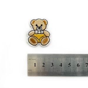 Puede incluir: Un pequeño parche bordado para planchar que representa un oso de peluche de dibujos animados con un mono amarillo. El oso es marrón con detalles blancos y sostiene un pequeño corazón blanco.