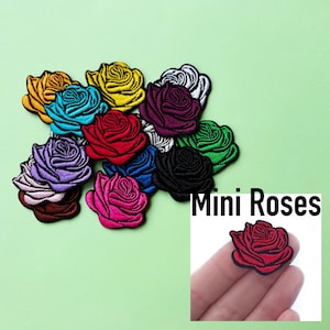 以下が含まれることがあります： さまざまな色のバラが描かれた、12個のミニアイロン接着パッチのセット。パッチは円形に配置され、中央には「Mini Roses」の文字が書かれている。1つのパッチが指に貼られている