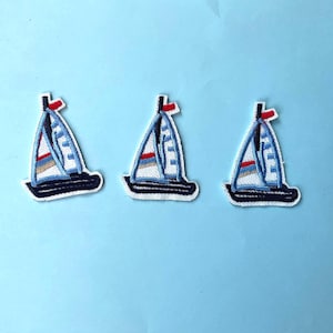 Op de afbeelding: Drie geborduurde zeilbootpatches. Elke patch heeft een wit zeil met blauwe en rode accenten, een marineblauwe romp en een rode vlag bovenaan de mast. De patches liggen op een lichtblauwe achtergrond.