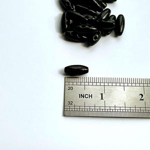 Peut inclure: Plusieurs perles en bois noires de forme ovale, d'environ 2,5 cm de long, sont présentées sur un fond blanc. Une règle est placée à côté d'une perle pour l'échelle. Les perles ont une finition brillante et sont probablement destinées à l'artisanat.