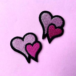 Può includere: Due toppe ricamate a forma di cuore in tonalità di rosa, con un contorno nero. I cuori sono sovrapposti, con un cuore più piccolo, rosa scuro, racchiuso in un cuore più grande, rosa chiaro. Le toppe sono su uno sfondo rosa.