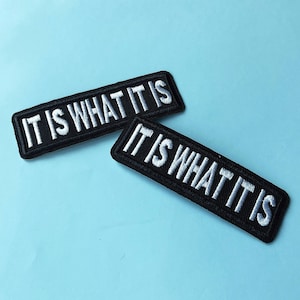 Könnte beinhalten: Zwei schwarze, rechteckige, gestickte Aufnäher mit dem Schriftzug "IT IS WHAT IT IS" in weißen Blockbuchstaben. Die Aufnäher haben einen schwarzen Rand und sind vor einem hellblauen Hintergrund platziert.