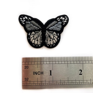 Könnte beinhalten: Ein gestickter Schmetterlings-Patch in Schwarz und Silber, etwa 2,5 cm breit. Der Patch wird neben einem Lineal zur Größenreferenz gezeigt.