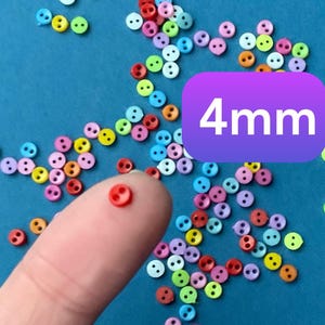 Könnte beinhalten: Nahaufnahme einer Fingerspitze, die einen kleinen roten Knopf hält. Der Knopf hat einen Durchmesser von 4 mm. Der Hintergrund ist eine blaue Oberfläche, die mit vielen kleinen, bunten Knöpfen bedeckt ist.